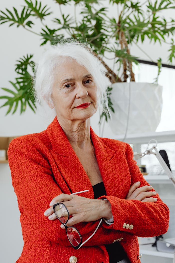 Senior woman in a red blazer confidently standing with arms crossed in an office setting.