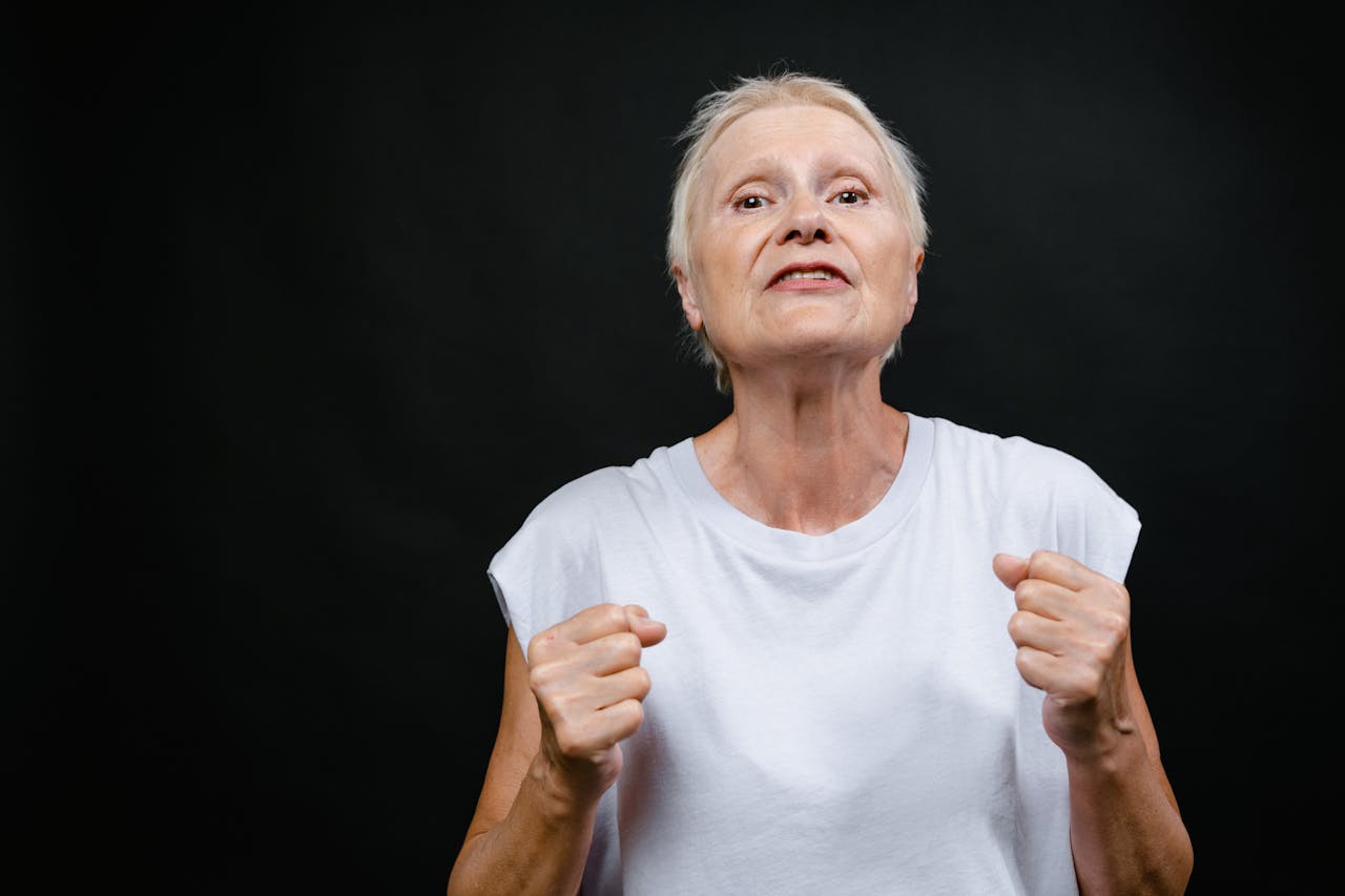 Determined elderly woman in white shirt clenching fists against black background.